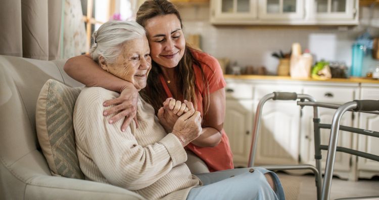 A woman embraces her elderly relative who is sitting on the couch.