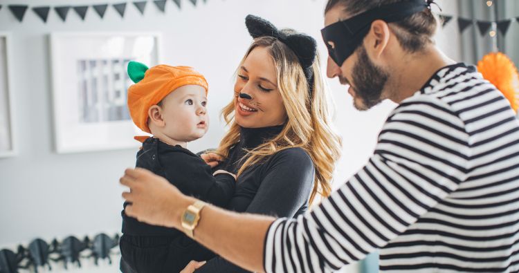A mother in a cat costumer carves a pumpkin with her child in a bat costume.
