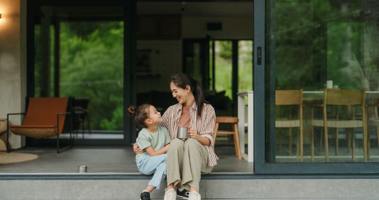 A mom sits on her stoop drinking a cup of coffee with her young daughter.