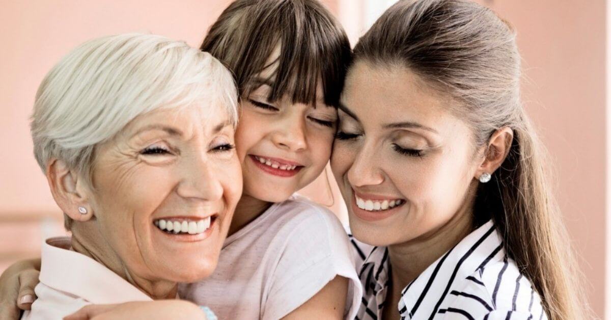 A grandmother, mother, and young daughter smile while embracing tightly