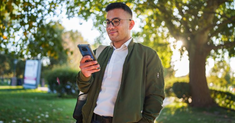 A young man smiling at his phone while standing outdoors in a park