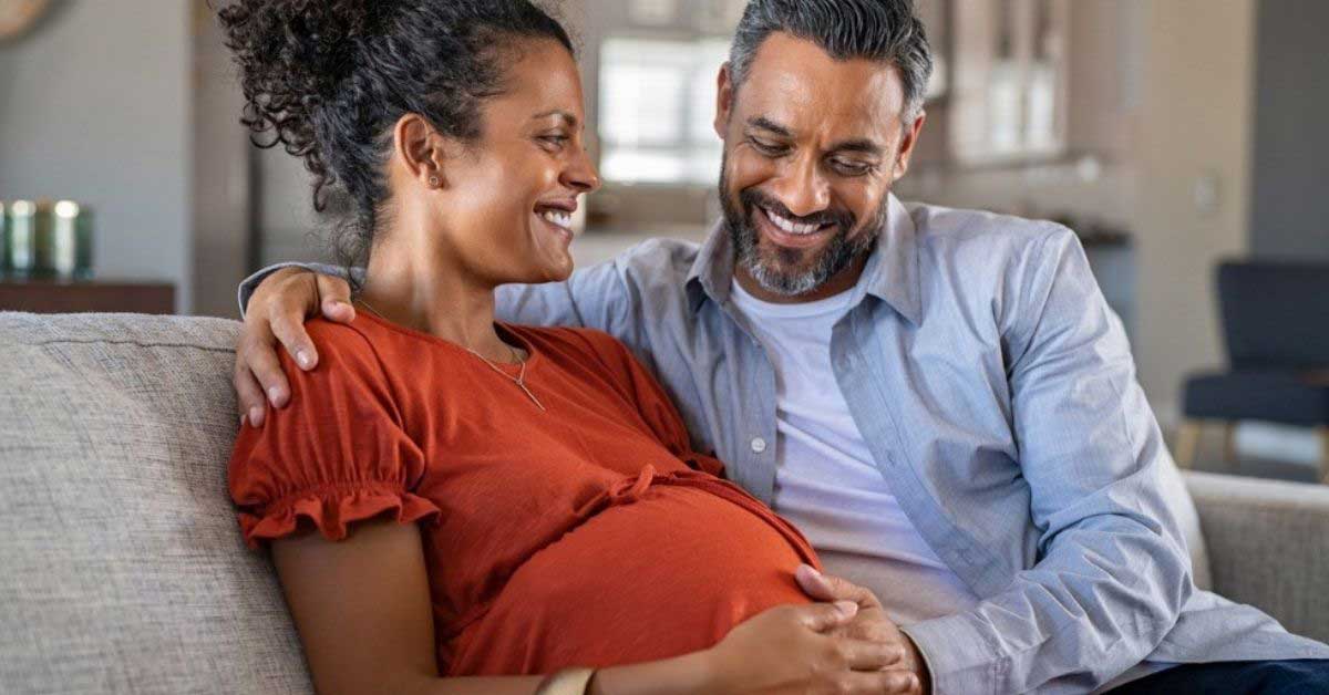 A young couple sit on a couch looking lovingly at the woman's pregnant belly