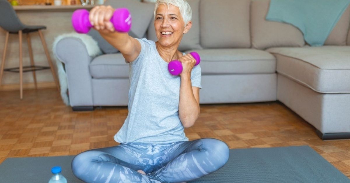 A women lifting weights while sitting on a yoga mat.