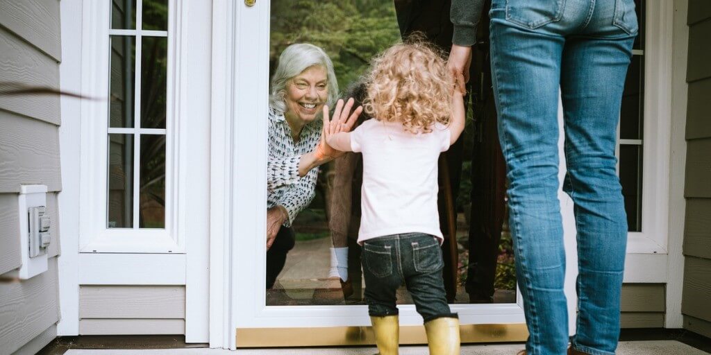 A grandmother puts her hand on a glass door to say hello to her grandchild for safety