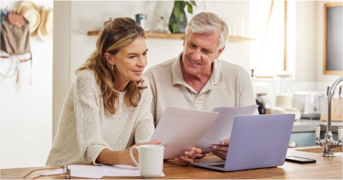 A retired couple sit in their kitchen reviewing their retirement plan on a laptop.