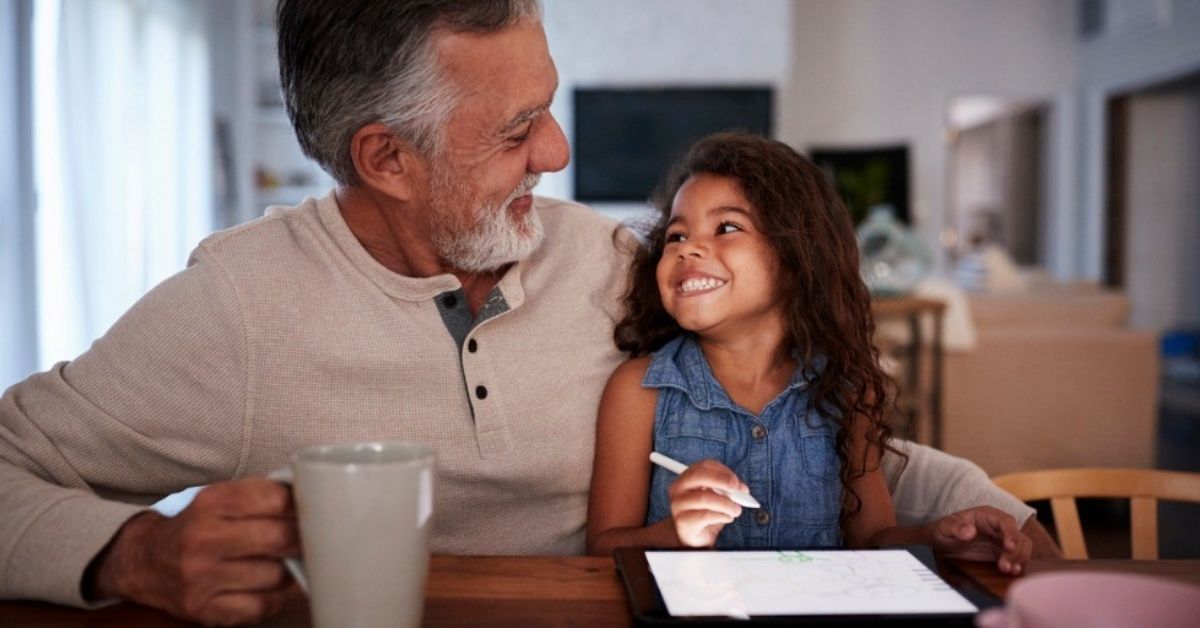 An older man sits with his granddaughter as she plays on an iPad