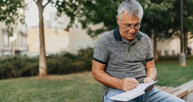 A man sits on a bench while writing in his notebook.