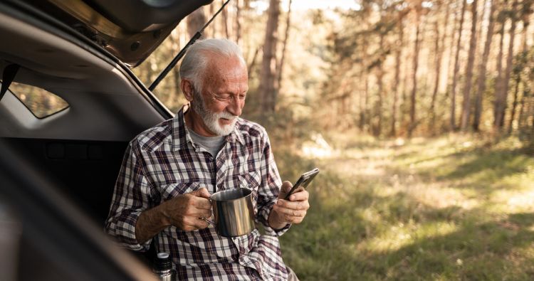 An older gentleman drinks coffee outside while looking at his phone.