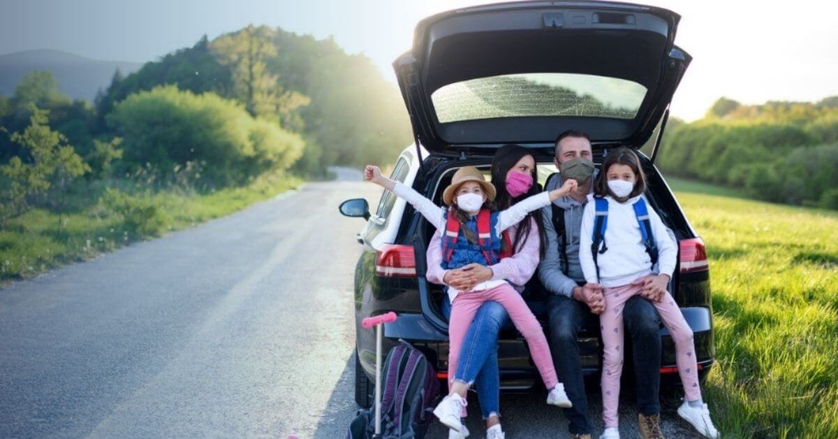 A young family sit in the back of their car wearing face masks during a road trip.