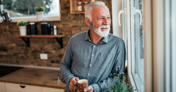 A man looks out his kitchen window while holding a cup of coffee.