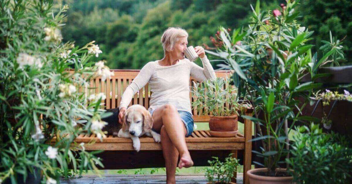 An older woman relaxes outside with a cup of coffee and her dog;.
